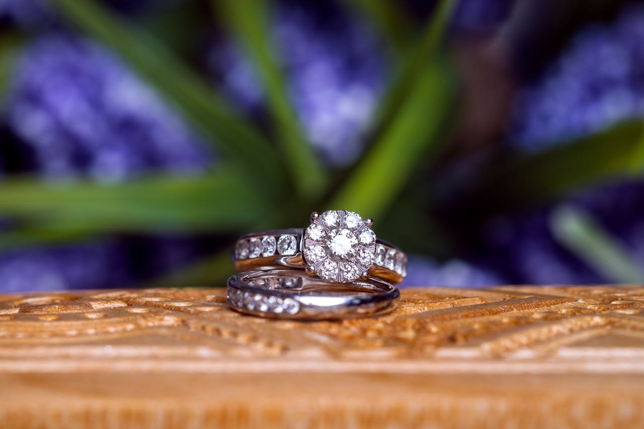 services-02 Close-up of diamond wedding rings on carved wood against a blurred floral background.