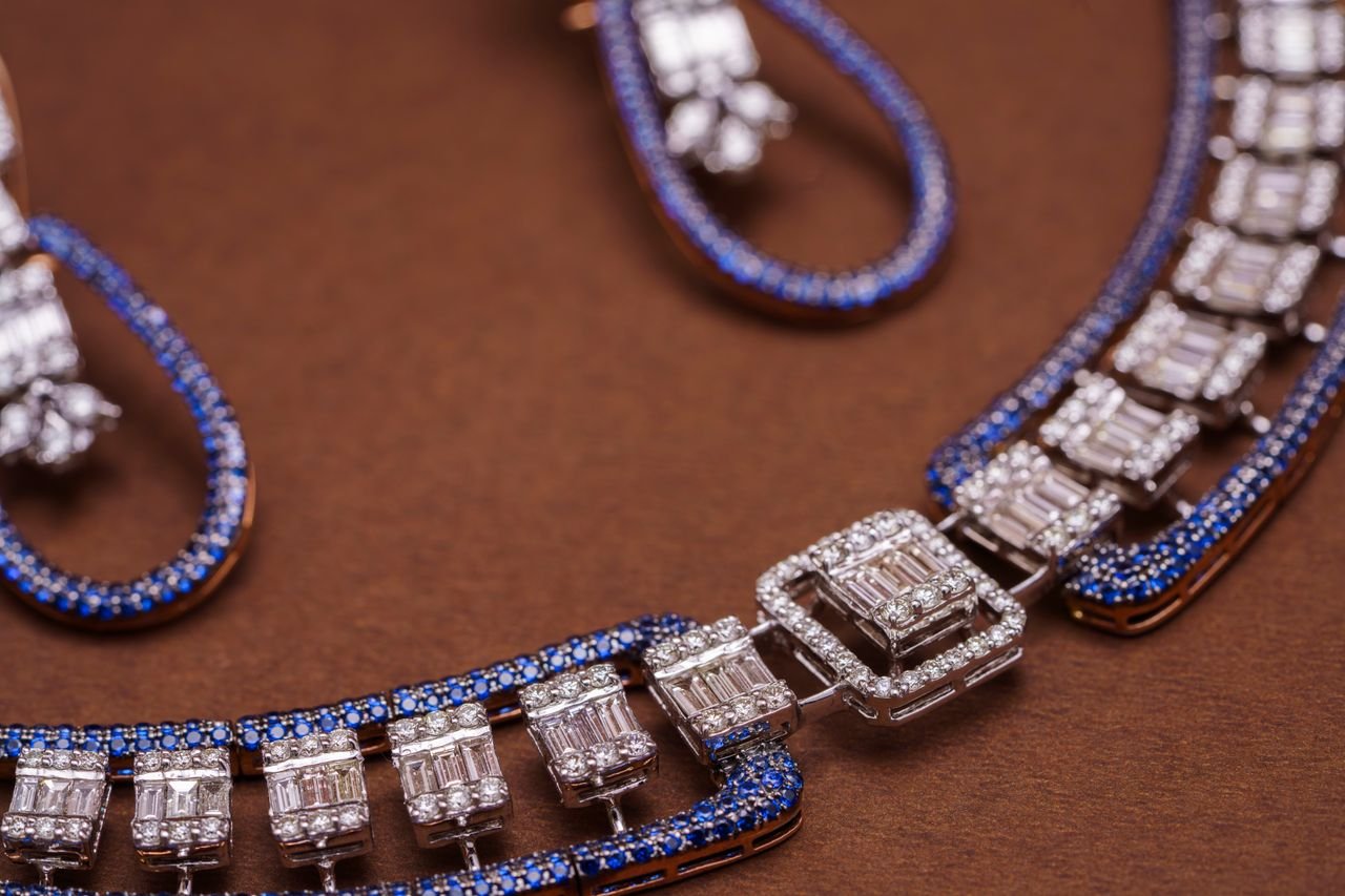 gallery-2 Close-up of an exquisite blue sapphire and diamond necklace and earrings against a brown background.